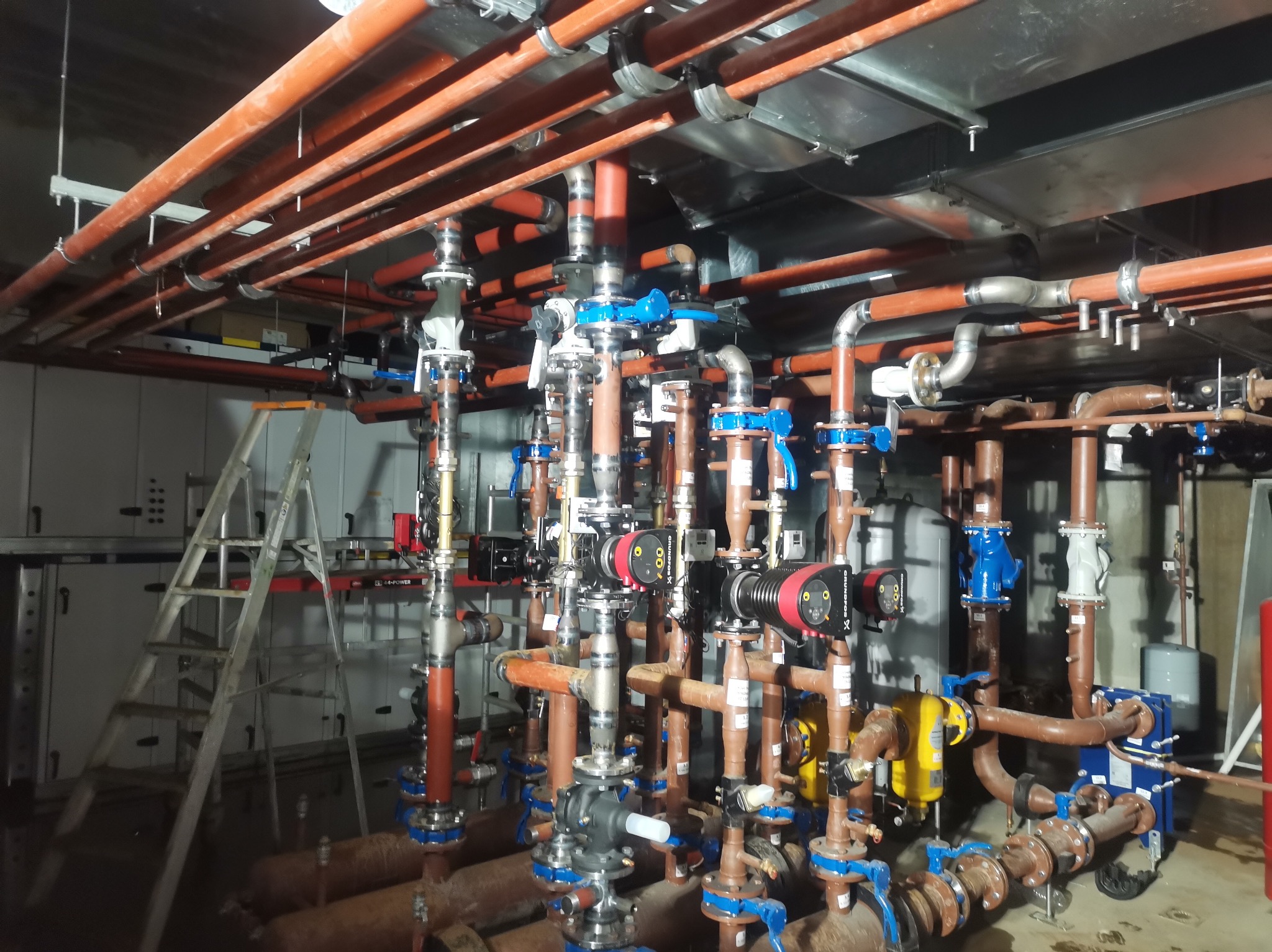 Rozbudowana instalacja rur miedzianych z zaworami i pompami w kotłowni, widoczna drabina aluminiowa i szafy sterownicze na tle.