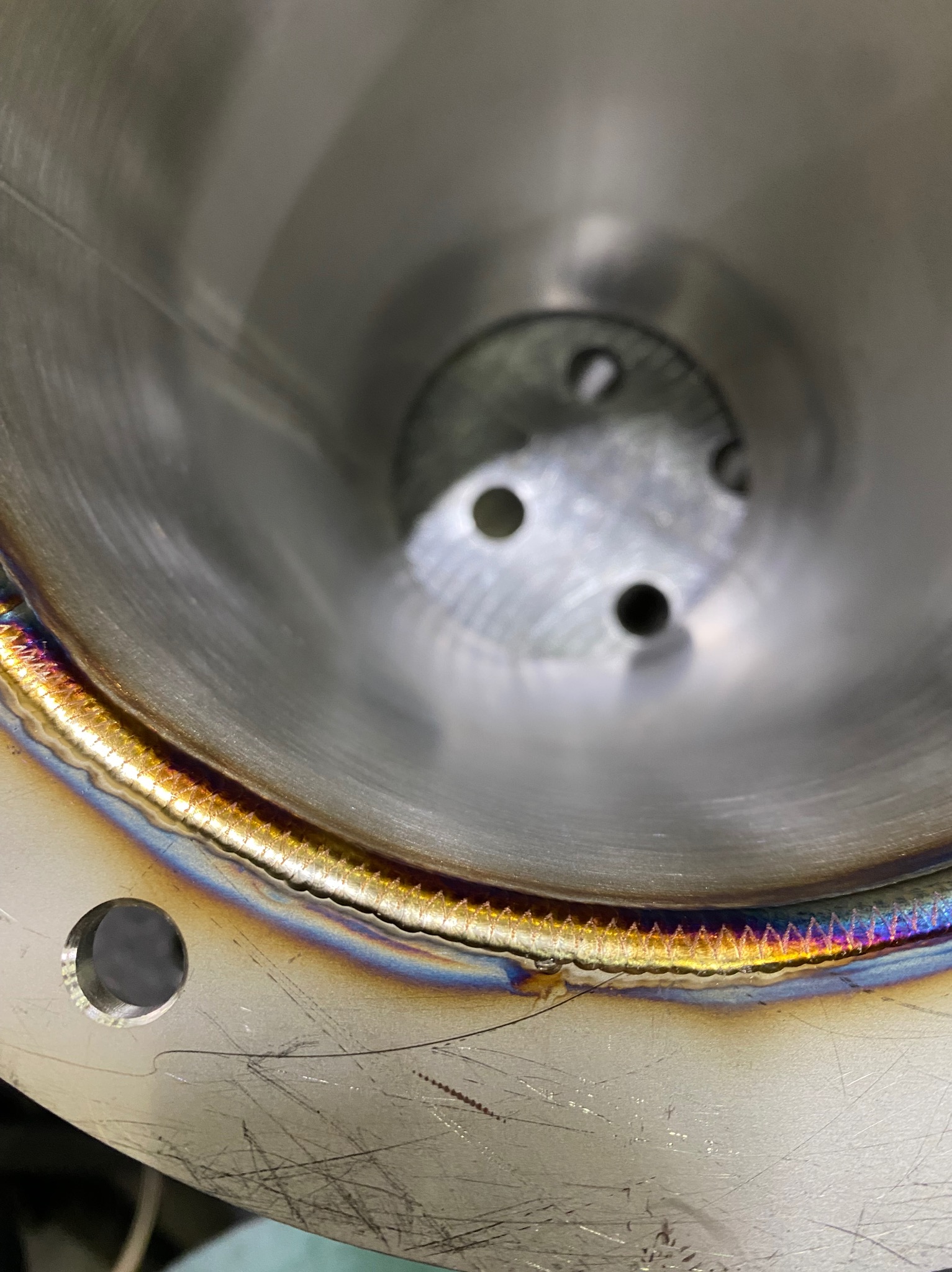 Wnętrze metalowej rury z widocznym, precyzyjnym spoiwem spawalniczym o tęczowych przebarwieniach. Na dnie widoczne otwory montażowe.