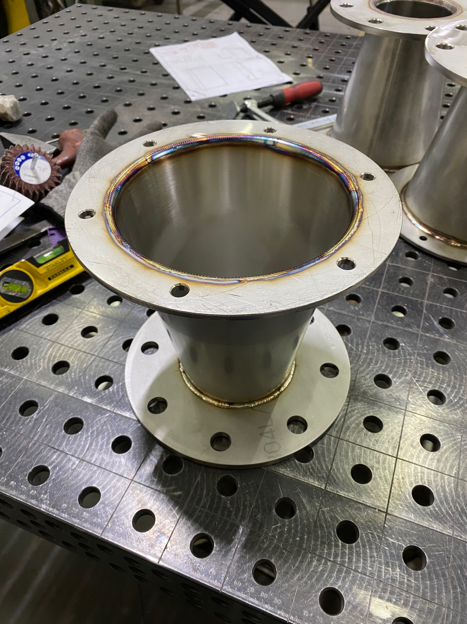 Element konstrukcyjny ze stali nierdzewnej z widocznymi spawami, leżący na perforowanym stole warsztatowym. Widoczne narzędzia i dokumentacja obok.