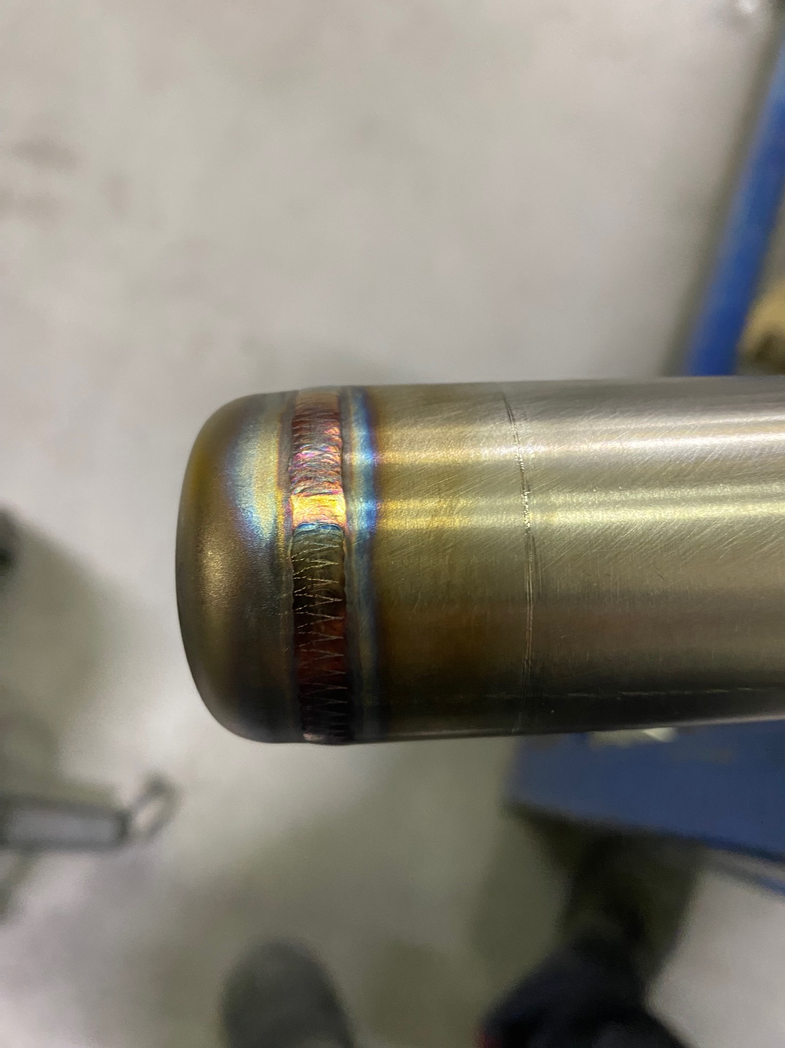 Detalne zbliżenie na spawany element ze stali nierdzewnej, z widocznymi kolorowymi przebarwieniami termicznymi w miejscu łączenia. Widoczne ślady obróbki.