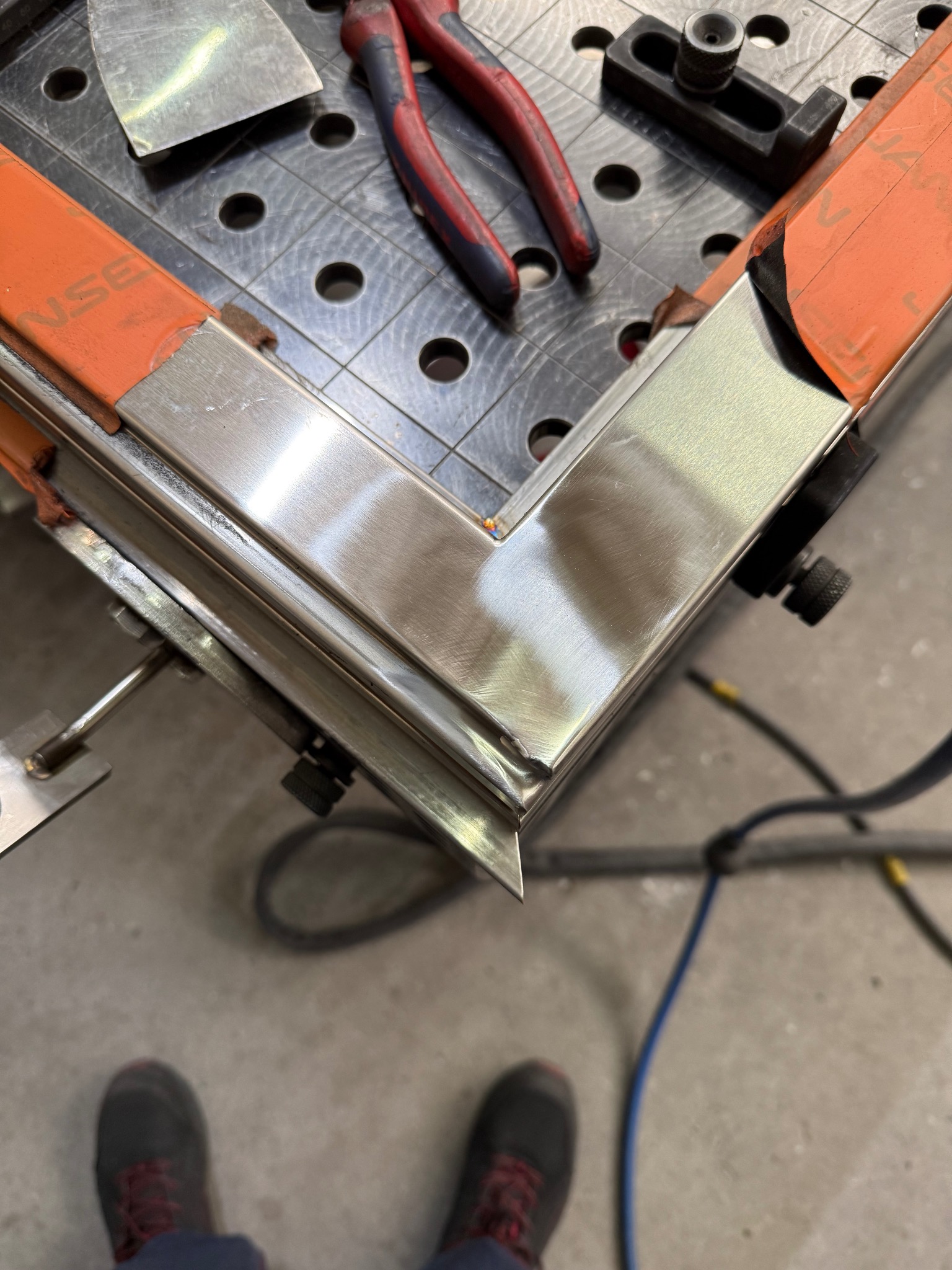 Spawany narożnik ramy ze stali nierdzewnej na stole spawalniczym z narzędziami. Widoczne ślady polerowania i mocowania. Precyzyjne wykonanie.