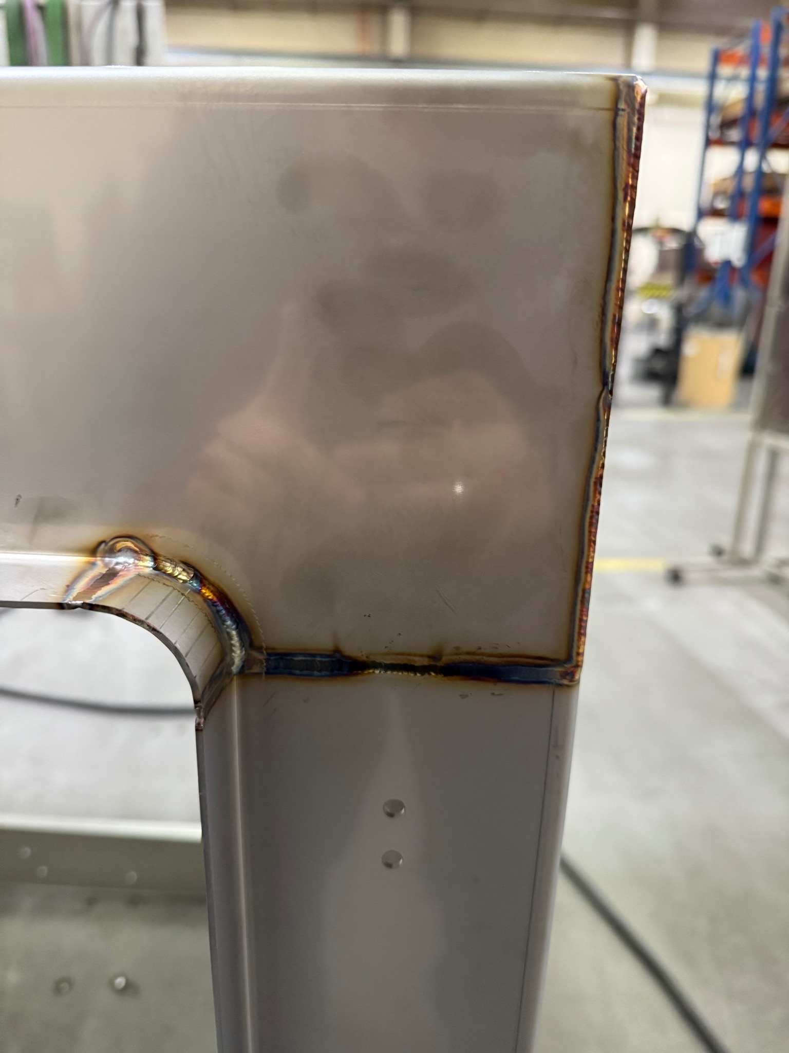 Szczegółowe zbliżenie na spawany narożnik metalowej konstrukcji. Widoczne precyzyjne spawy i opalizujące zabarwienie metalu w strefie spawania. Tło rozmyte, industrialne.