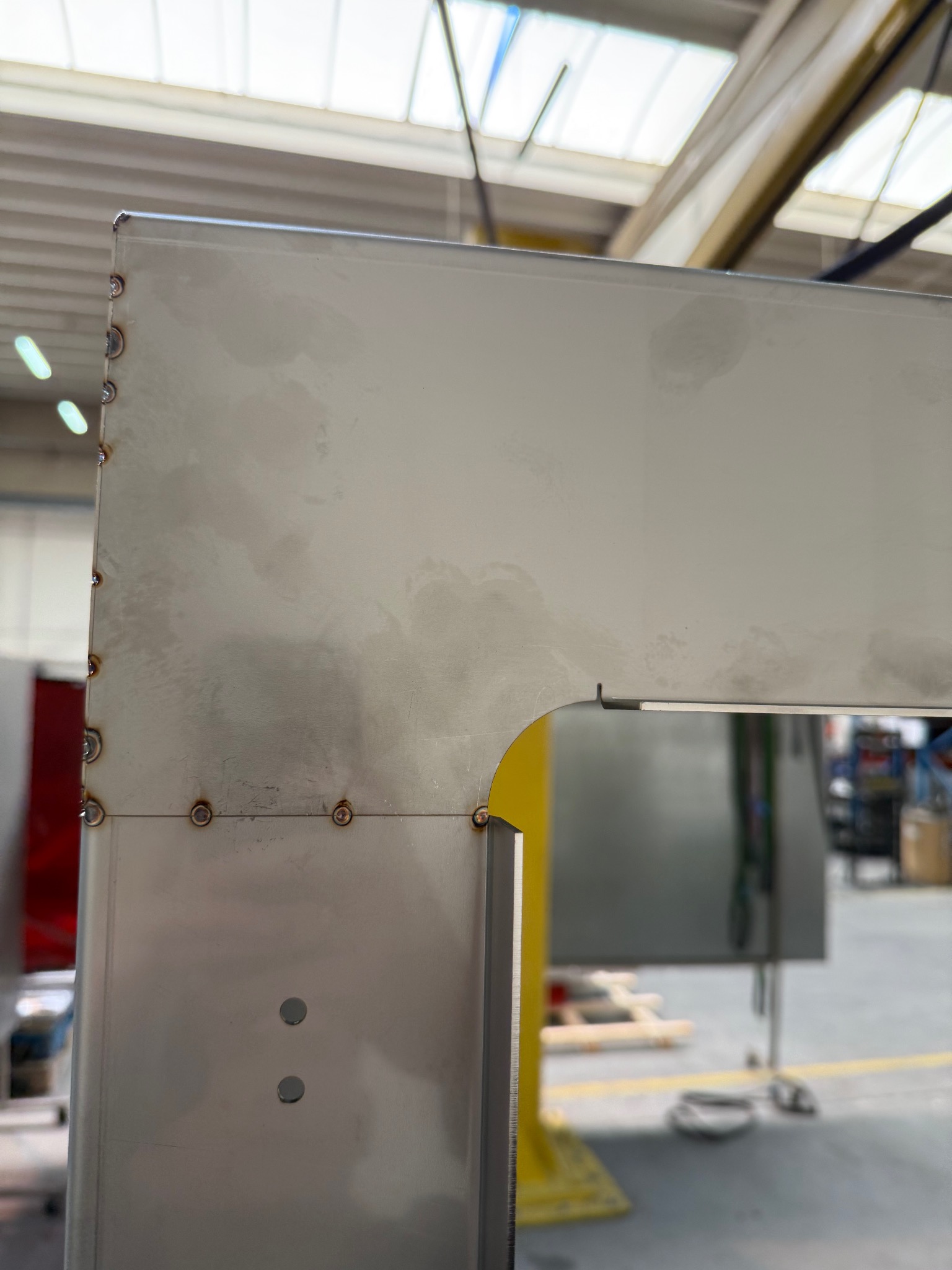 Metalowa konstrukcja spawana, widoczne spawy punktowe i gładka powierzchnia. Fragment hali w tle, industrialny charakter.