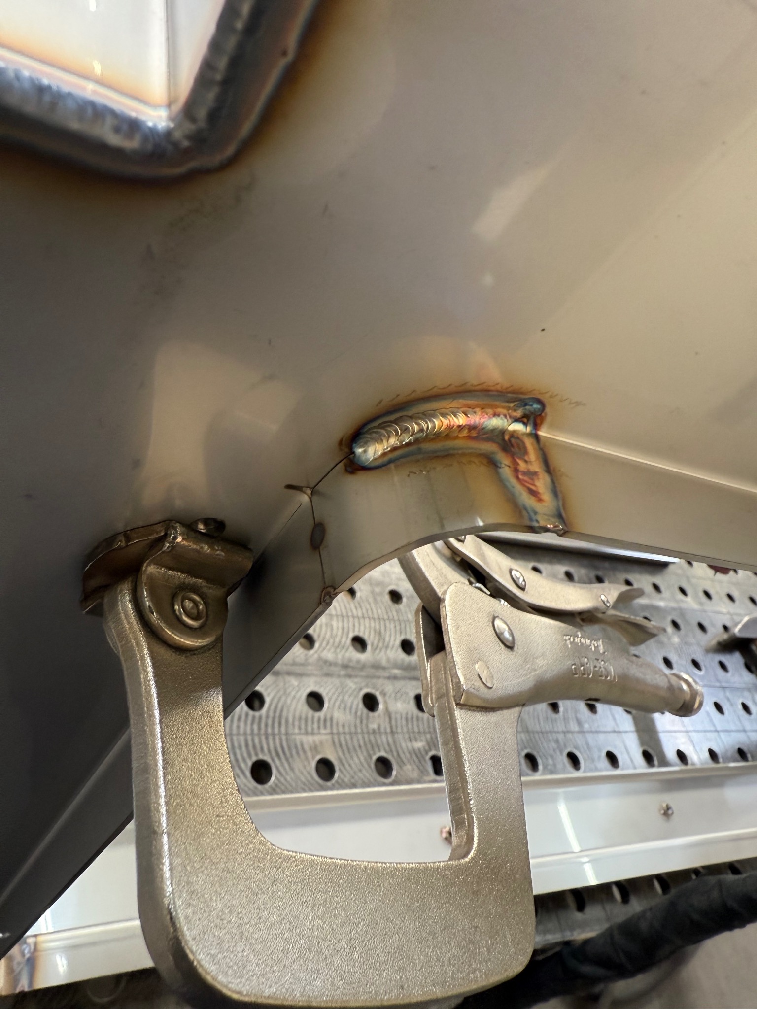 Metalowy element mocowany ściskiem spawalniczym, widoczny spaw z charakterystycznym zabarwieniem, tło stanowi stół spawalniczy z otworami.