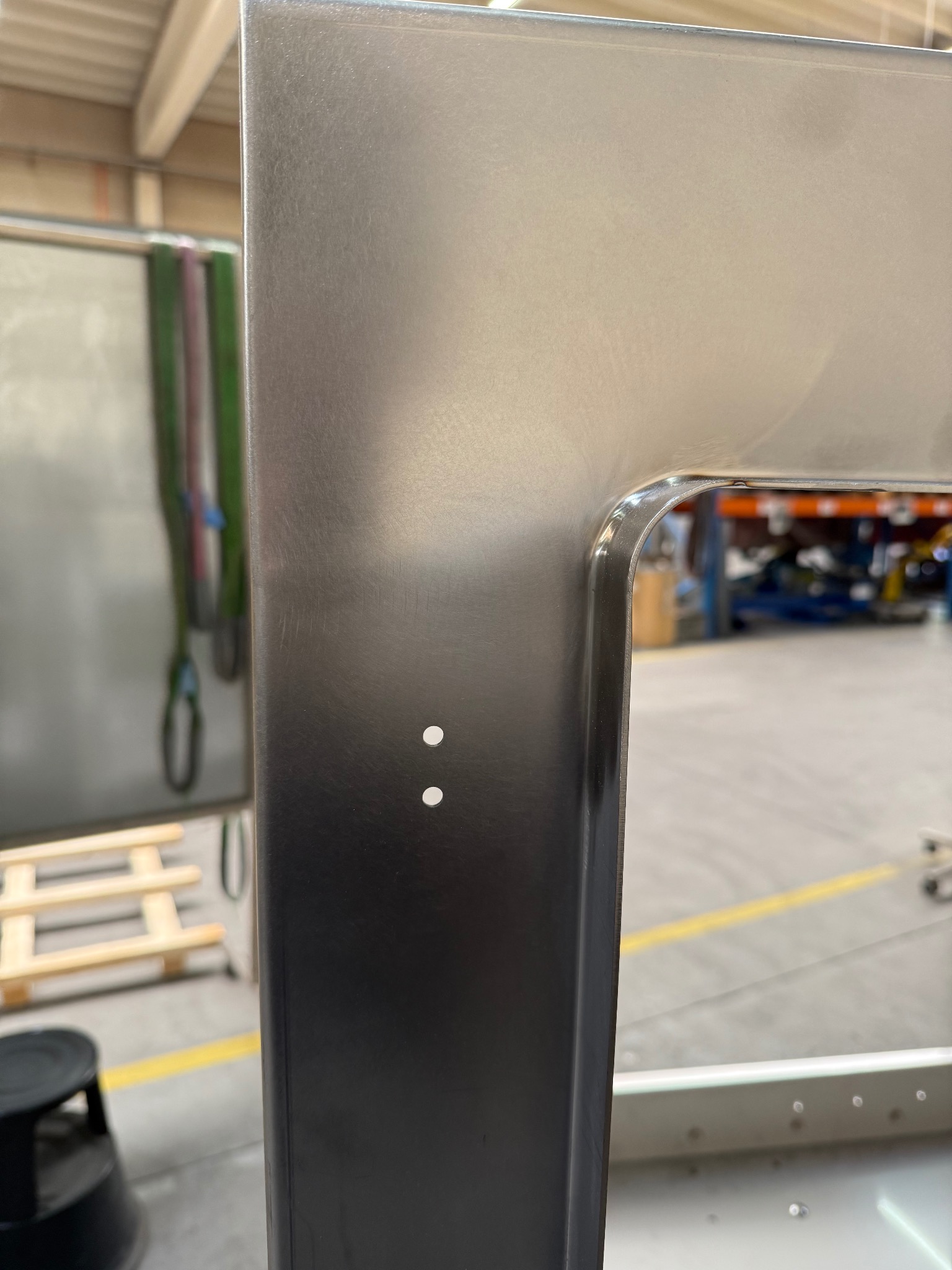 Detal wykończenia narożnika ramy z polerowanej stali nierdzewnej, z widocznymi dwoma otworami montażowymi i delikatną krzywizną krawędzi. Tło warsztatowe.