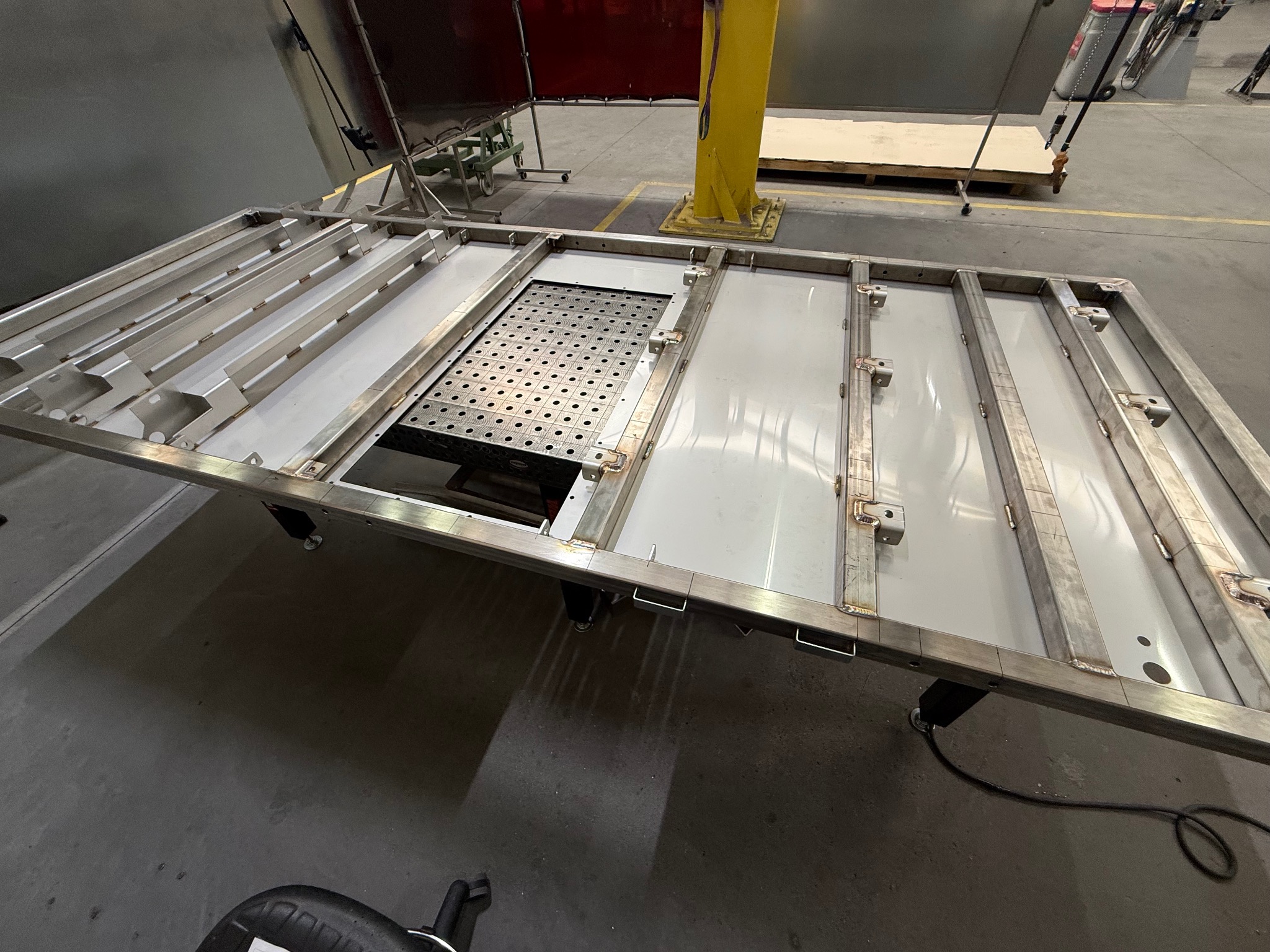 Metalowa konstrukcja z elementami spawanymi, prawdopodobnie rama lub podstawa, w hali produkcyjnej. Widoczne ślady spawania i elementy mocujące.