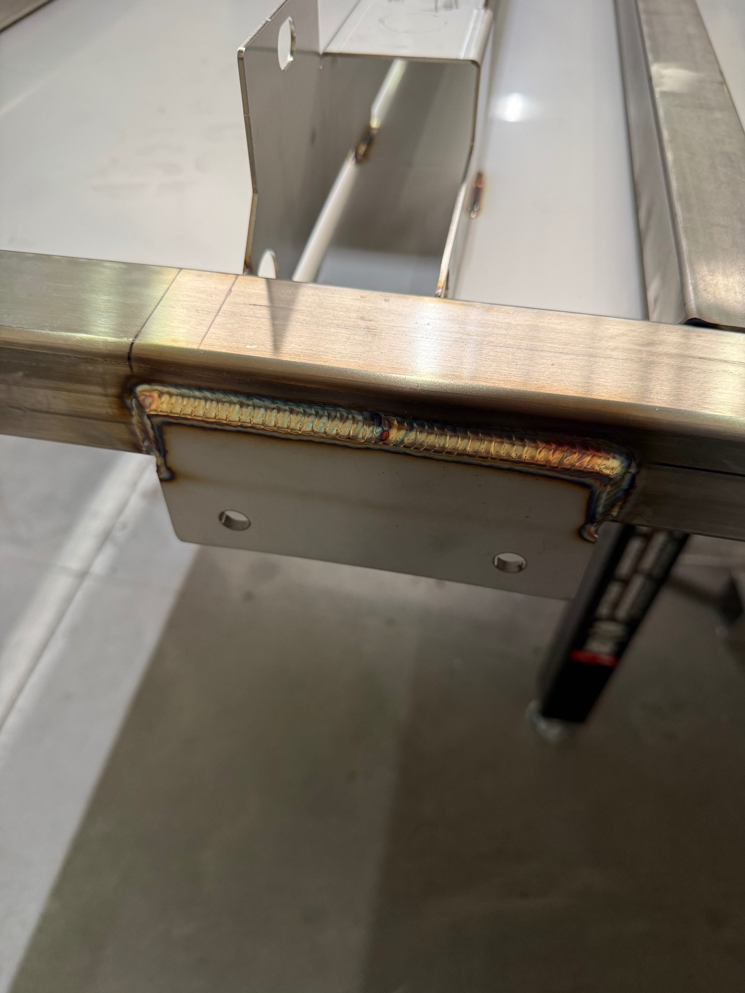 Szlifowany profil z metalu, spawany z płaskownikiem. Widoczne ślady po spawaniu i obróbce. Element konstrukcyjny z otworami. Jasne tło, zbliżenie na detal.