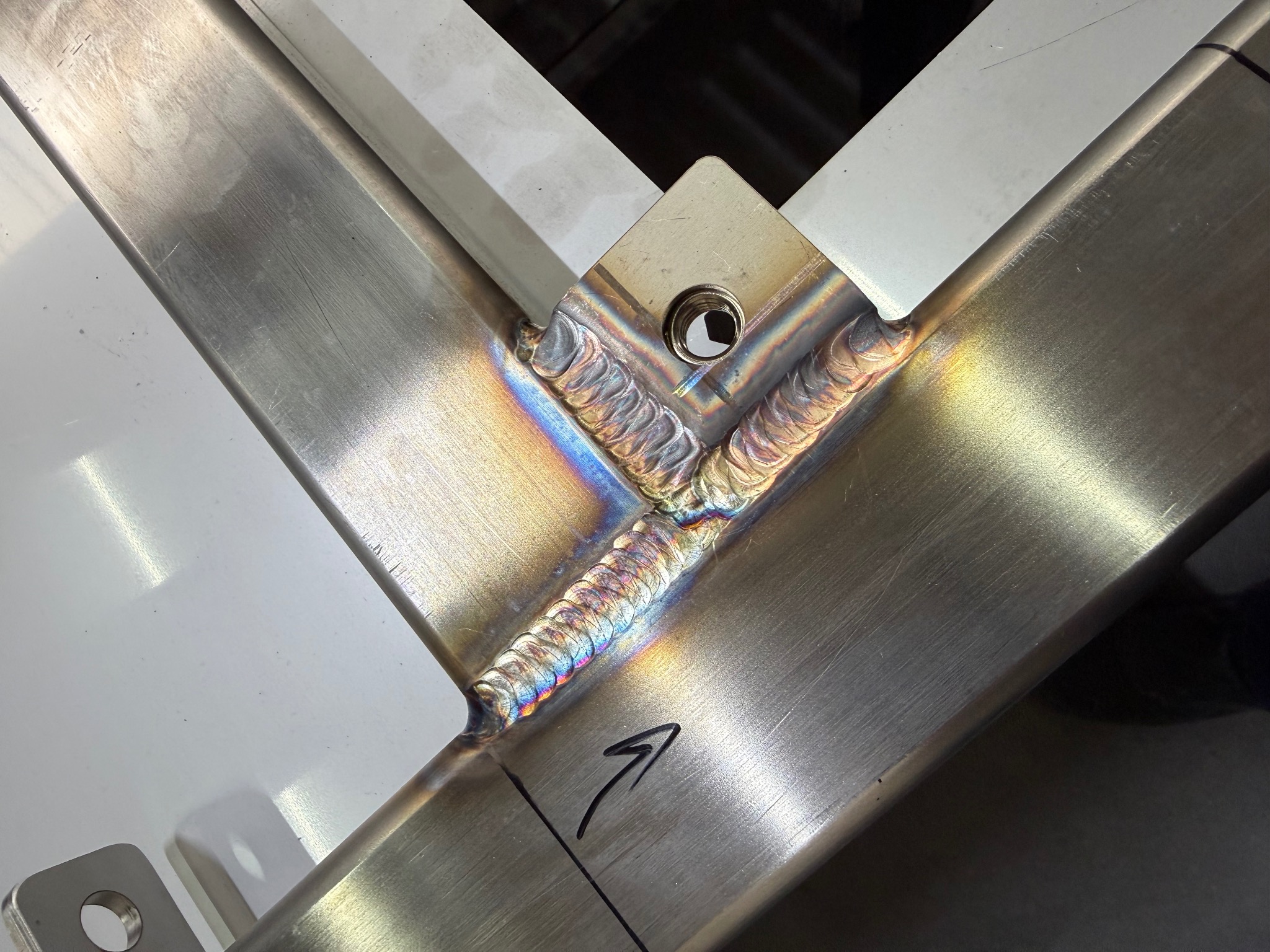 Szczegółowe zbliżenie na precyzyjne spawy łączące elementy metalowej konstrukcji. Widoczne tęczowe przebarwienia po spawaniu i czarna strzałka narysowana markerem.