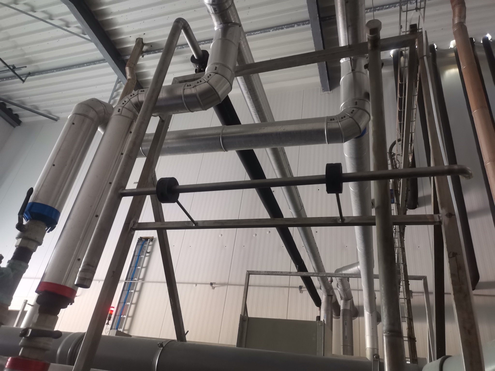 Kompleksowa instalacja przemysłowa z rurami o różnych średnicach, mocowana do sufitu. Widoczne elementy mocujące i fragment drabiny w tle.