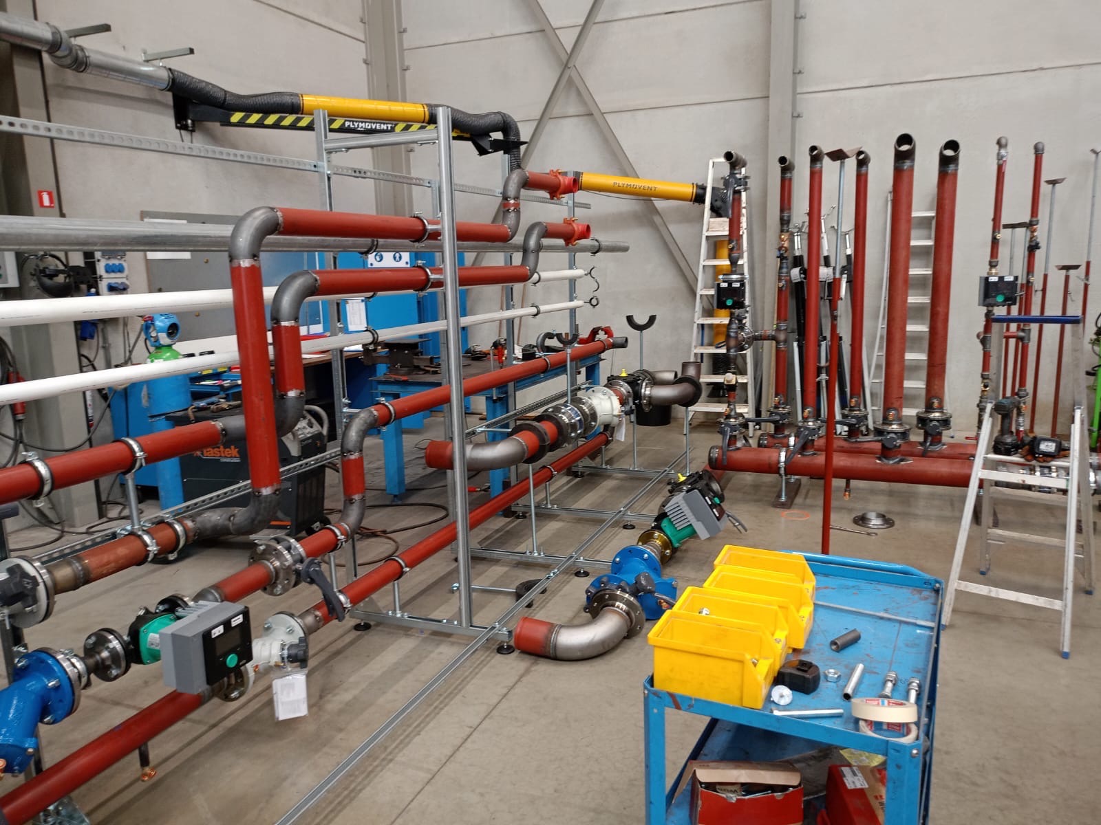 Rozbudowana instalacja przemysłowa z rur w kolorach czerwonym, szarym i żółtym, zamontowana w hali. Widoczne liczne złącza, zawory i elementy mocujące. Na pierwszym planie wózek narzędziowy.