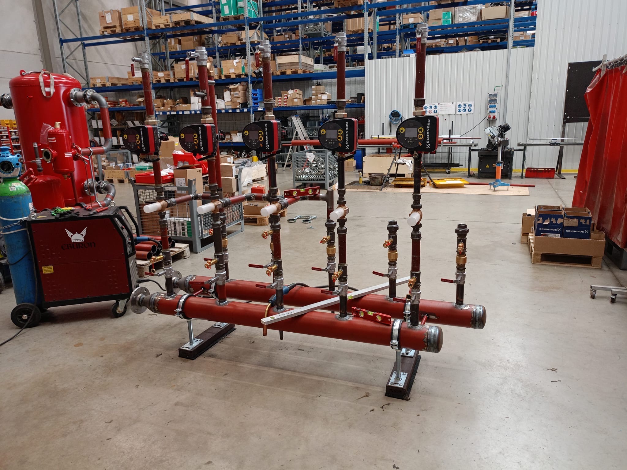 Czerwone rury z zaworami i pompami Grundfos X na metalowej konstrukcji w hali produkcyjnej, z widocznym sprzętem spawalniczym i regałami magazynowymi w tle.
