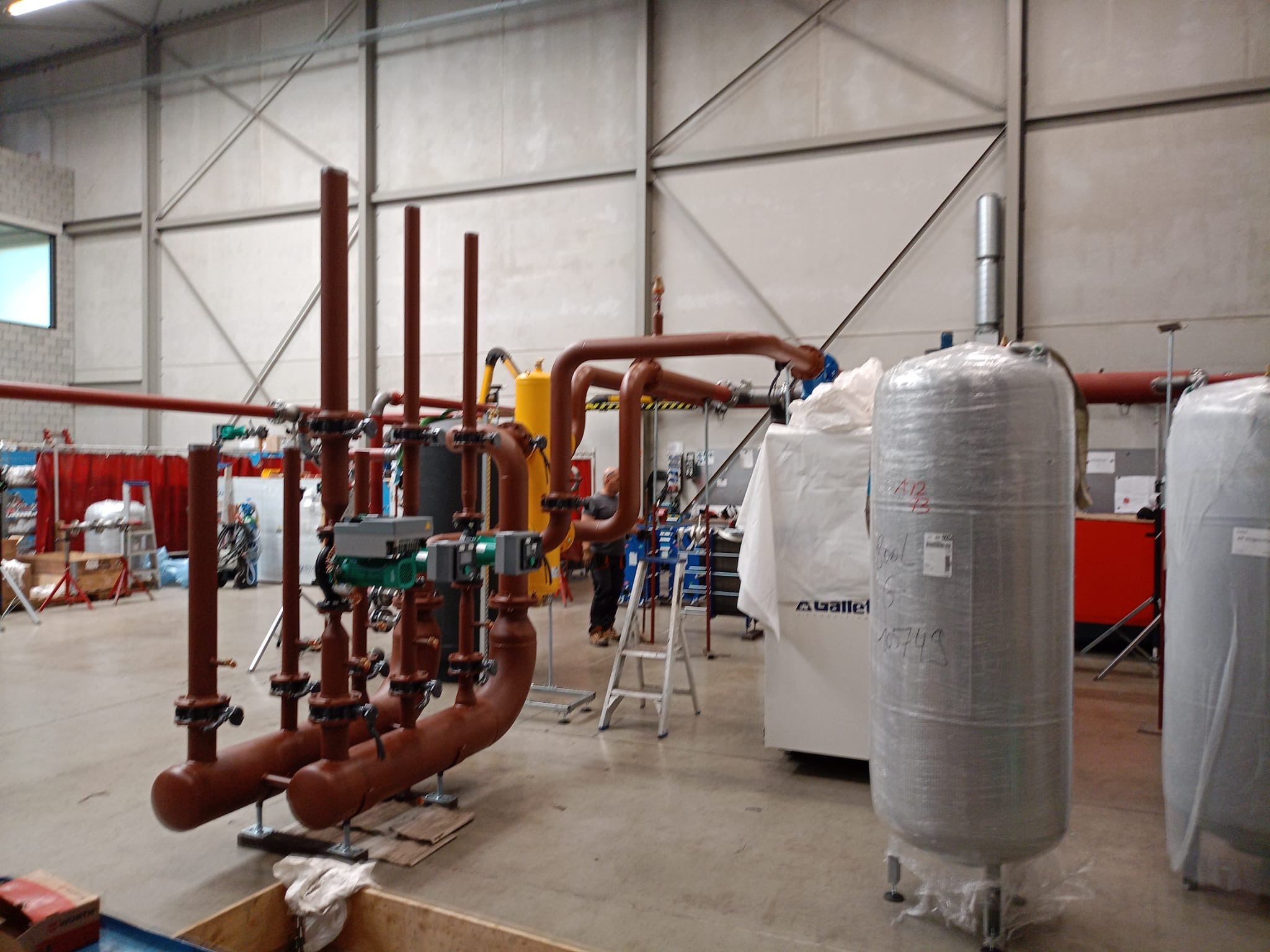 Brązowe rury przemysłowe z zaworami i zielonymi regulatorami w hali produkcyjnej. W tle zbiorniki owinięte folią i drabina. Widoczny fragment pracownika.
