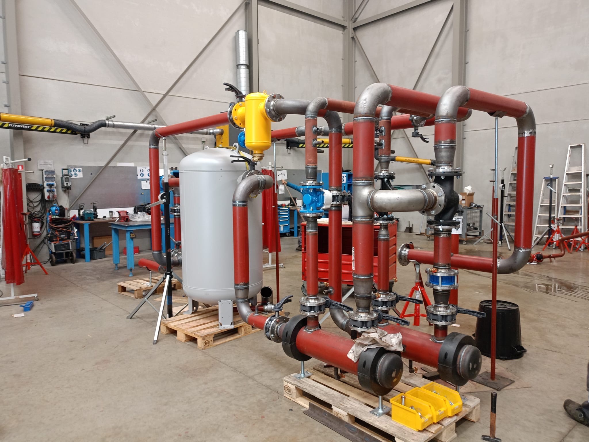 Konstrukcja z rur stalowych, czerwonych i szarych, z elementami żółtymi i niebieskimi, na paletach w hali produkcyjnej. Widoczne spawane łączenia i elementy mocujące.