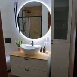 Nowoczesna łazienka z okrągłym lustrem z podświetleniem LED, białą umywalką nablatową na szafce z drewnianym blatem, czarną armaturą i kabiną prysznicową w tle, uzupełniona minimalistycznymi...