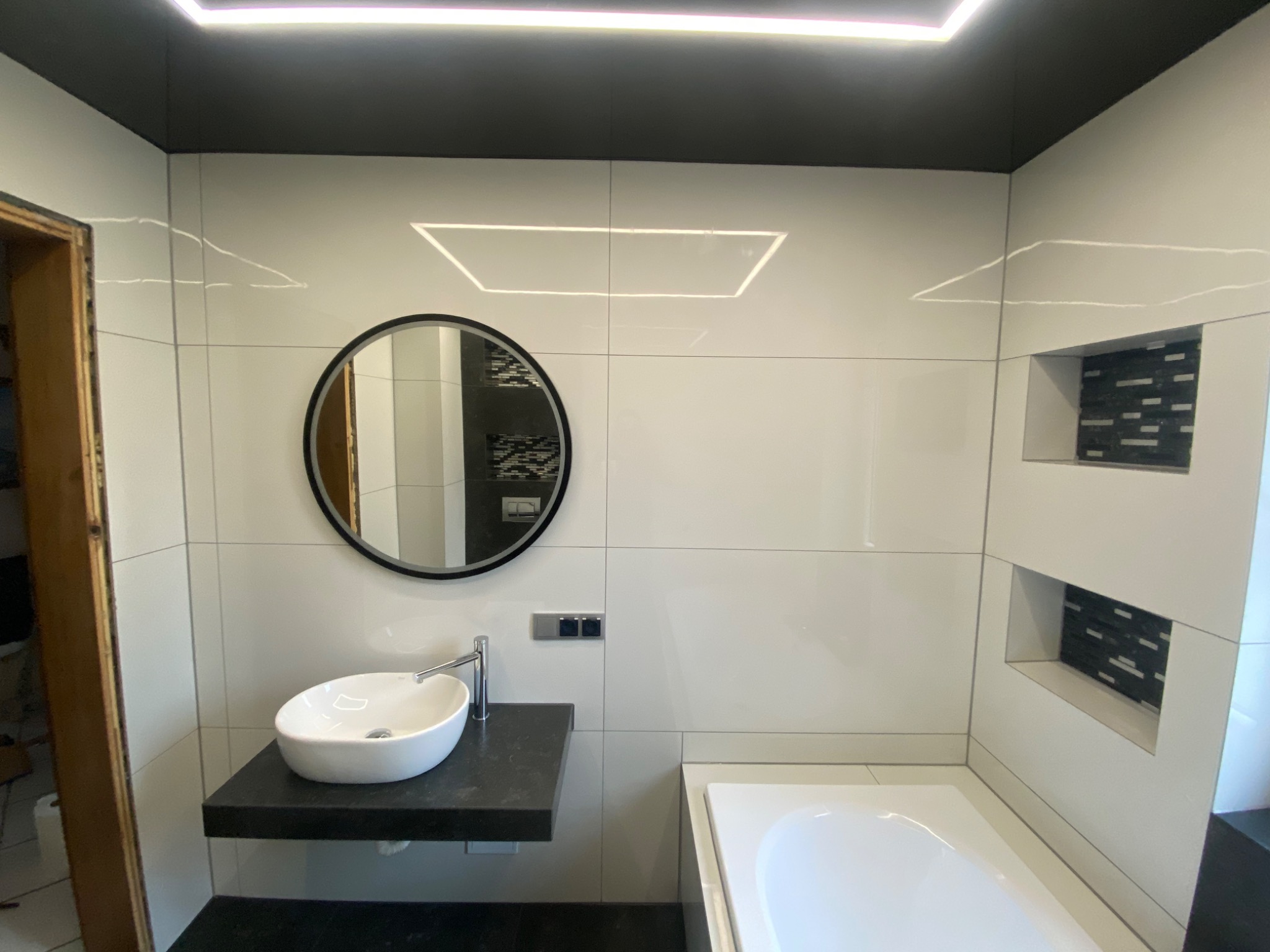 Nowoczesna łazienka z białymi płytkami, czarnym sufitem i blatem, okrągłym lustrem, oraz podświetleniem LED w suficie i wnęką z mozaiką.