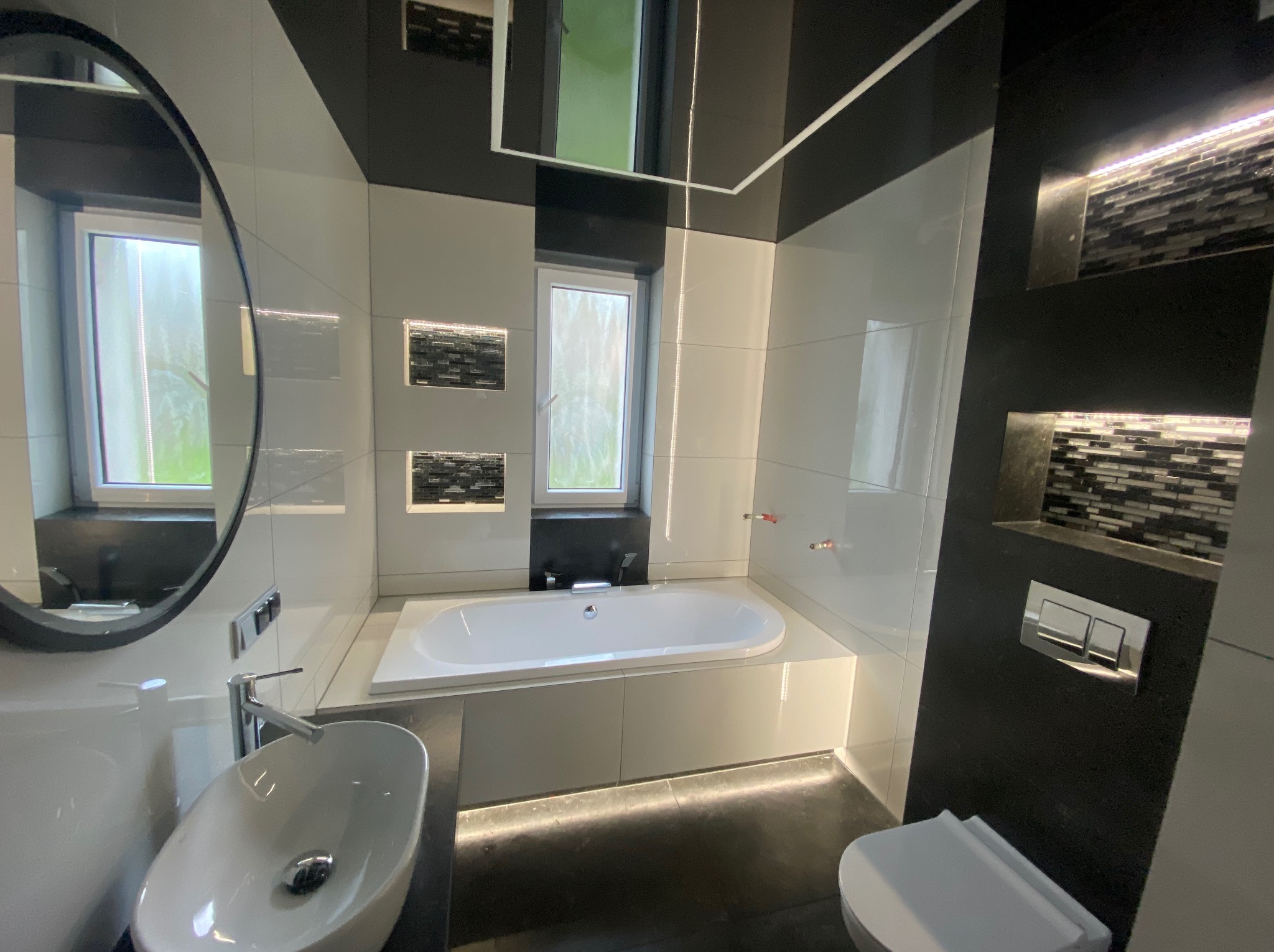 Nowoczesna łazienka w bieli i czerni z podświetleniem LED pod wanną, owalnym lustrem i umywalką nablatową.