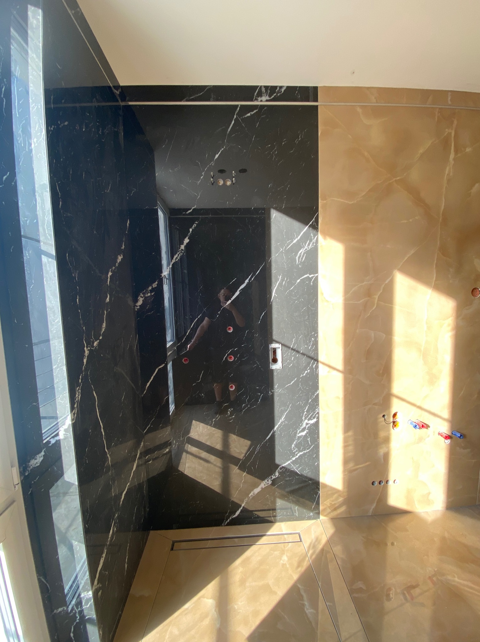 Nowoczesna łazienka w trakcie wykańczania z czarnymi i beżowymi płytkami imitującymi marmur, widoczne przyłącza wodne i odpływ liniowy w podłodze, oświetlona promieniami słonecznymi