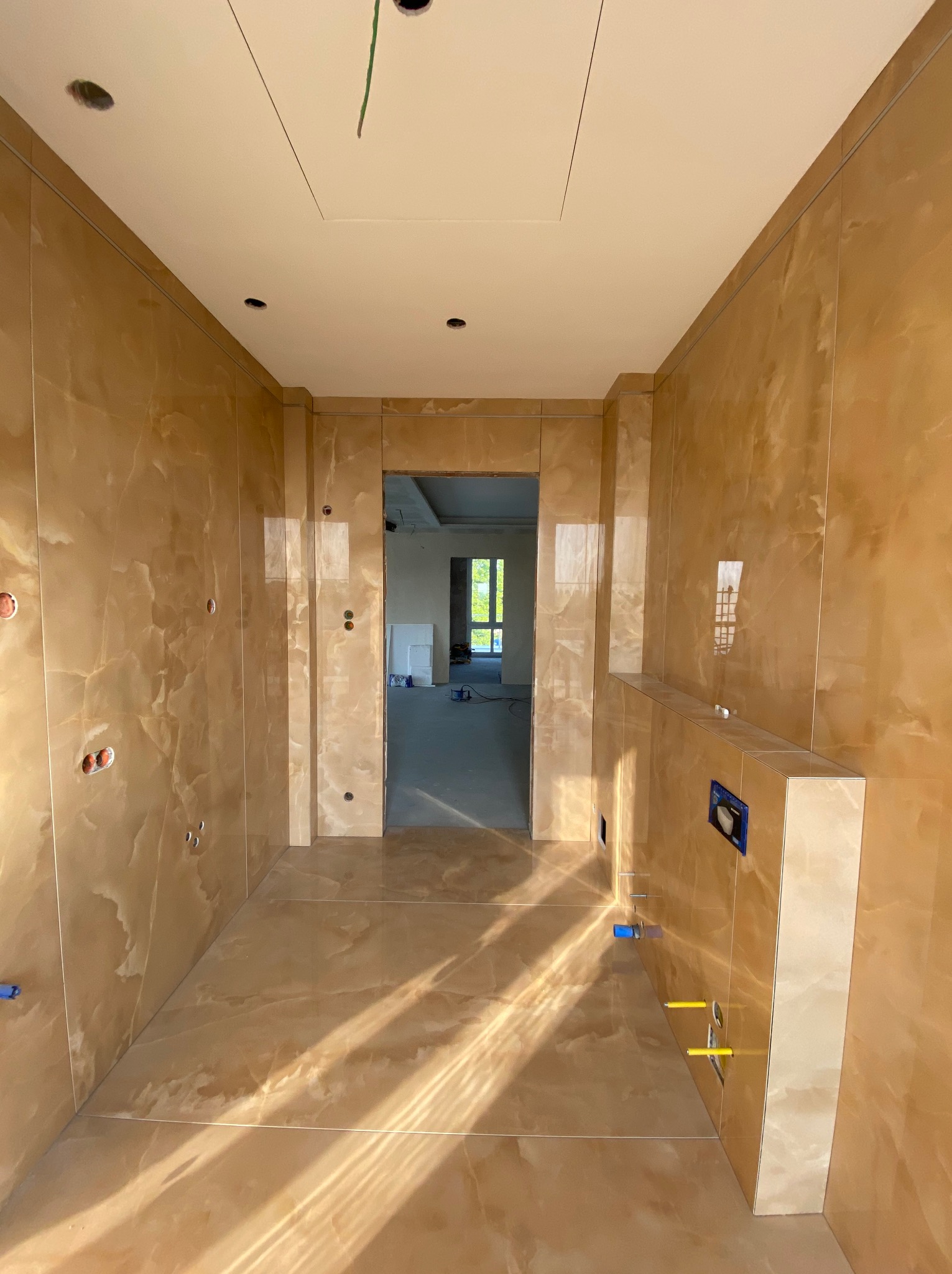 Wnętrze łazienki w trakcie remontu, wyłożone płytkami imitującymi marmur w odcieniach beżu i brązu, widoczne otwory na instalacje elektryczne i hydrauliczne, oraz stelaż podtynkowy WC z niebieskimi...