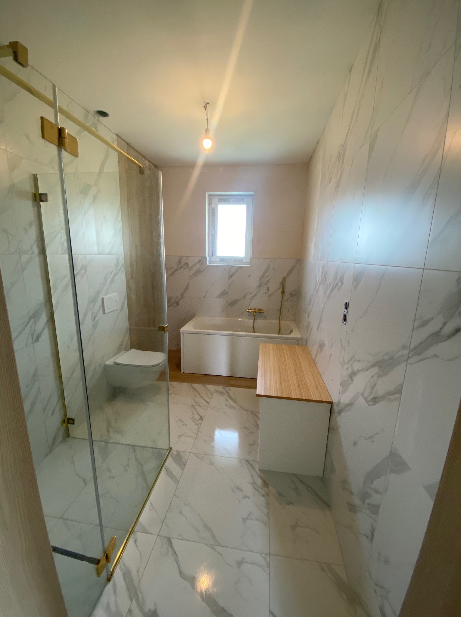 Wykończona łazienka z marmurowymi płytkami, kabiną prysznicową z złotymi elementami, wiszącą toaletą, białą wanną i drewnianym blatem.