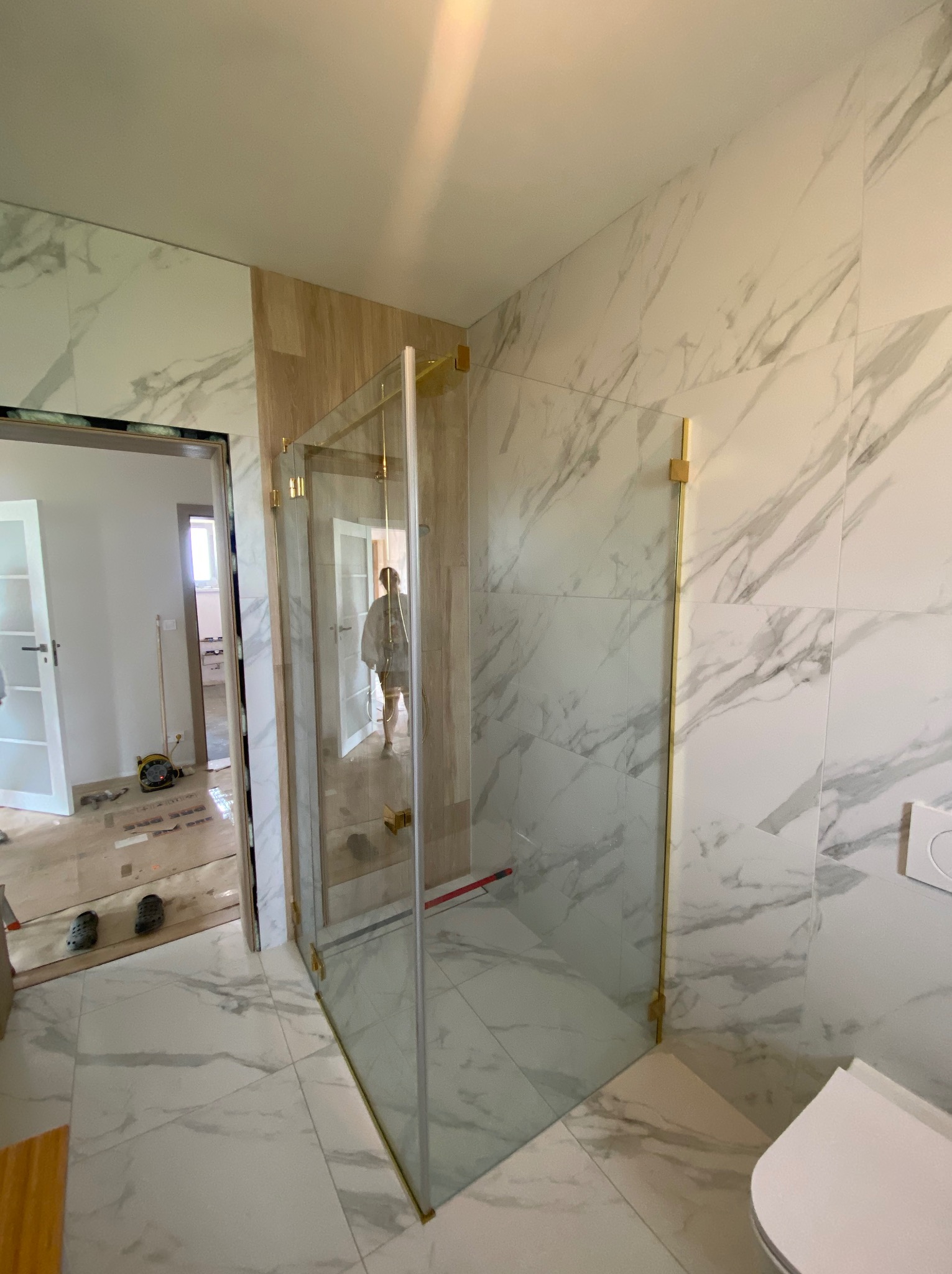 Nowoczesna kabina prysznicowa ze złotymi okuciami i marmurowymi płytkami w trakcie montażu, widoczne narzędzia i osoba w tle.