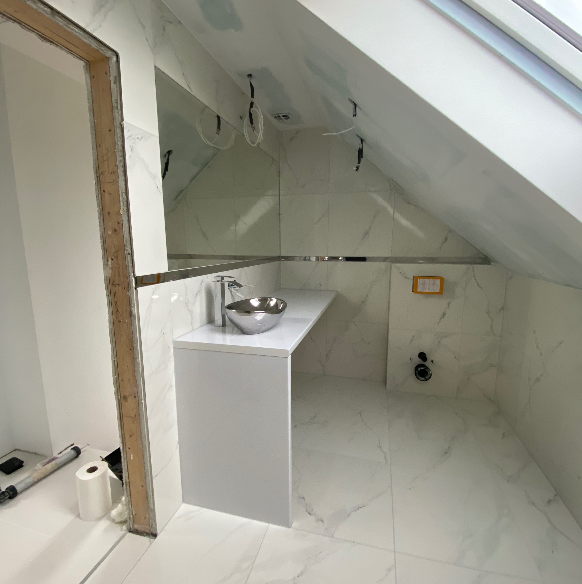 Łazienka w trakcie wykańczania na poddaszu: białe płytki imitujące marmur, srebrna umywalka nablatowa, niedokończone obramowanie drzwi i widoczne przewody elektryczne.