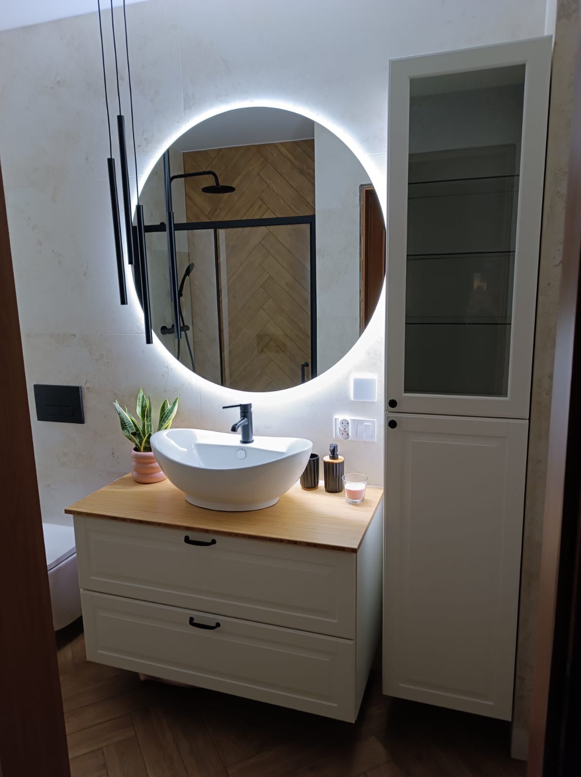 Nowoczesna łazienka z okrągłym lustrem z podświetleniem LED, białą umywalką nablatową na szafce z drewnianym blatem, czarną armaturą i kabiną prysznicową w tle, uzupełniona minimalistycznymi...