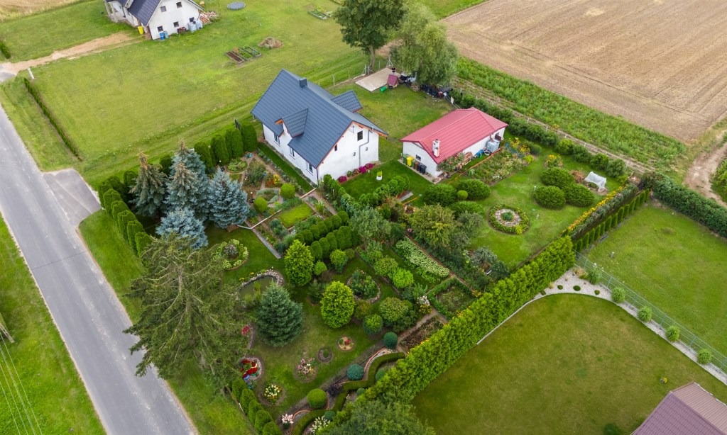 Widok z lotu ptaka na zadbany ogród z dwoma domami: jeden z szarym dachem, drugi z czerwonym, otoczony żywopłotem i różnorodną roślinnością.