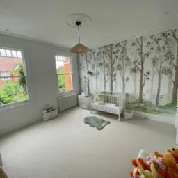 Jasny pokój dziecięcy z białymi ścianami, tapetą z motywem leśnym, białym łóżeczkiem i dywanikiem w kształcie misia, oświetlony lampą z abażurem z naturalnego materiału