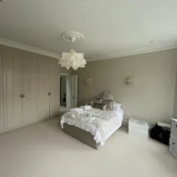 Neutralna sypialnia z beżowymi ścianami, zabudowaną szafą, teksturowanym dywanem i łóżkiem z dekoracyjnymi poduszkami oraz lampą w kształcie kwiatu.