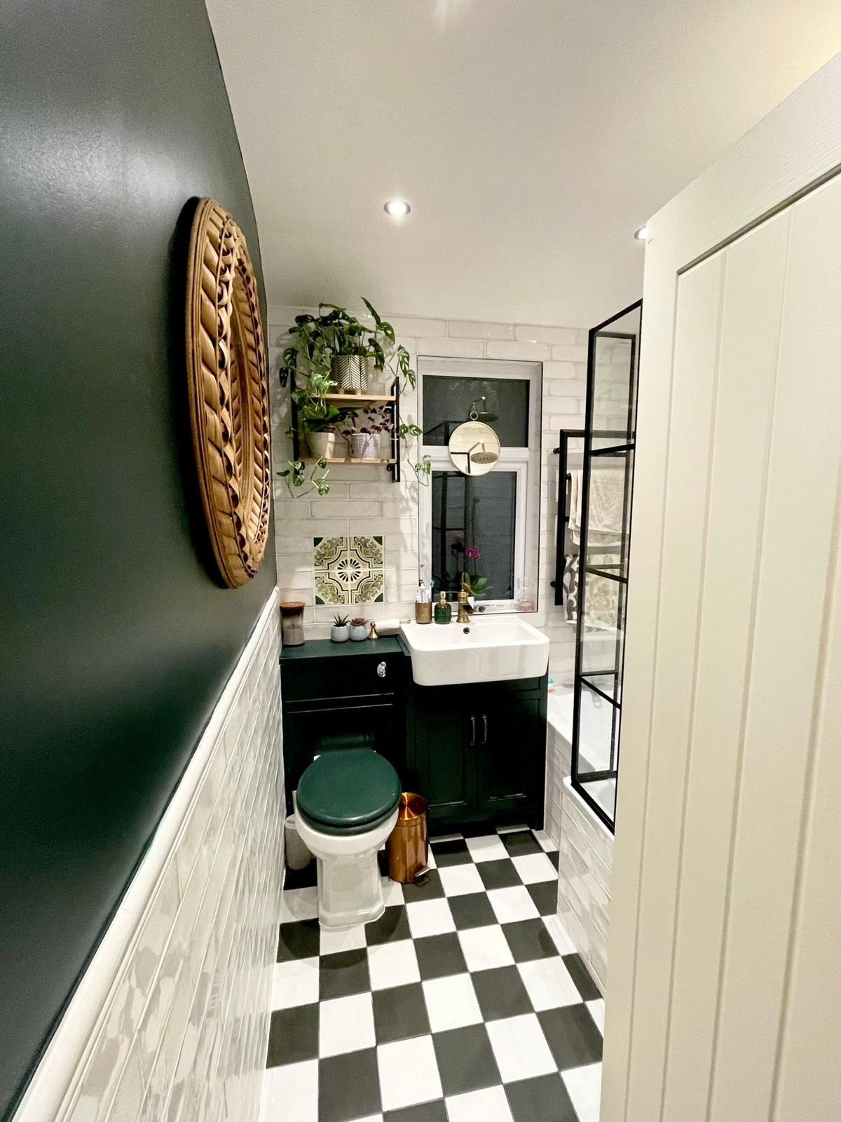 Nowoczesna łazienka z czarno-białą szachownicą na podłodze, ciemnozielonymi ścianami i szafką pod umywalką, dekoracyjnym lustrem na ścianie i roślinami doniczkowymi na półce.