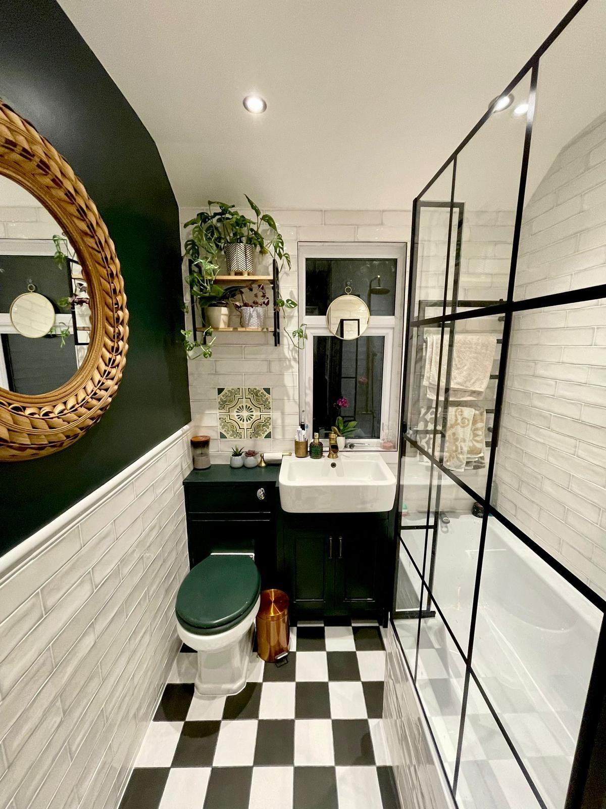 Ukończona łazienka w stylu retro z czarno-białą szachownicą na podłodze, ciemnozielonymi ścianami i szafką pod umywalką, oraz wiklinowym lustrem na ścianie.