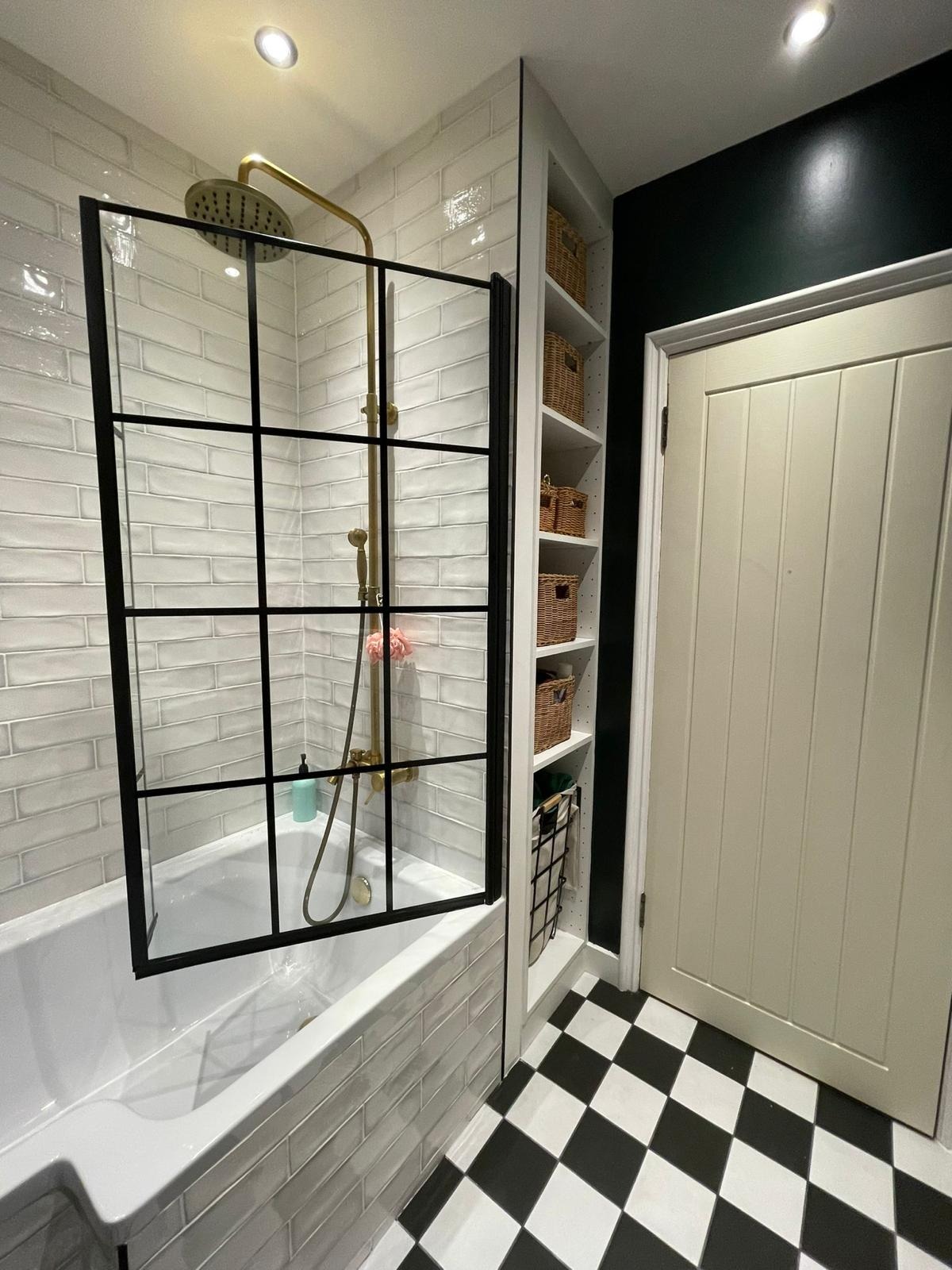 Biała łazienka z wanną, czarną ramą kabiny prysznicowej, złotą armaturą, półkami z koszykami i czarno-białą podłogą w szachownicę.