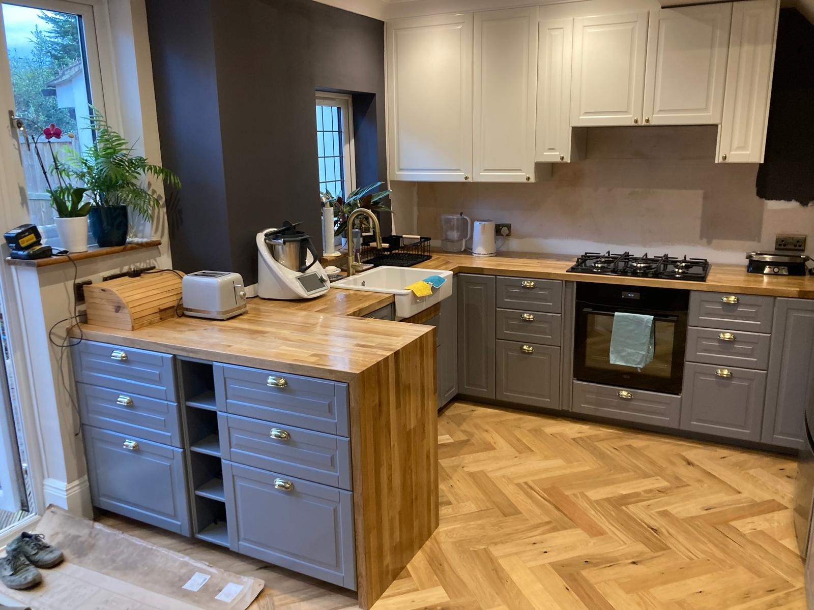 Szara zabudowa kuchenna z drewnianym blatem, białymi szafkami wiszącymi i parkietem w jodełkę; widoczne sprzęty AGD i rośliny doniczkowe.