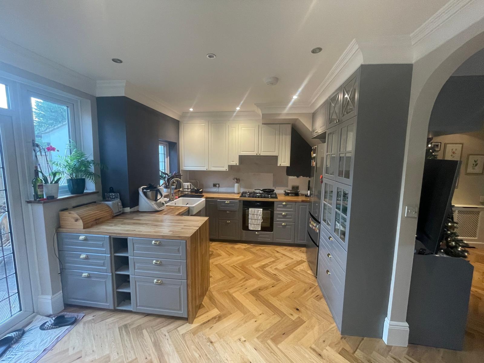 Jasna kuchnia po remoncie z szarymi szafkami dolnymi i białymi górnymi, drewnianym blatem i parkietem w jodełkę, widok na okno z roślinami.