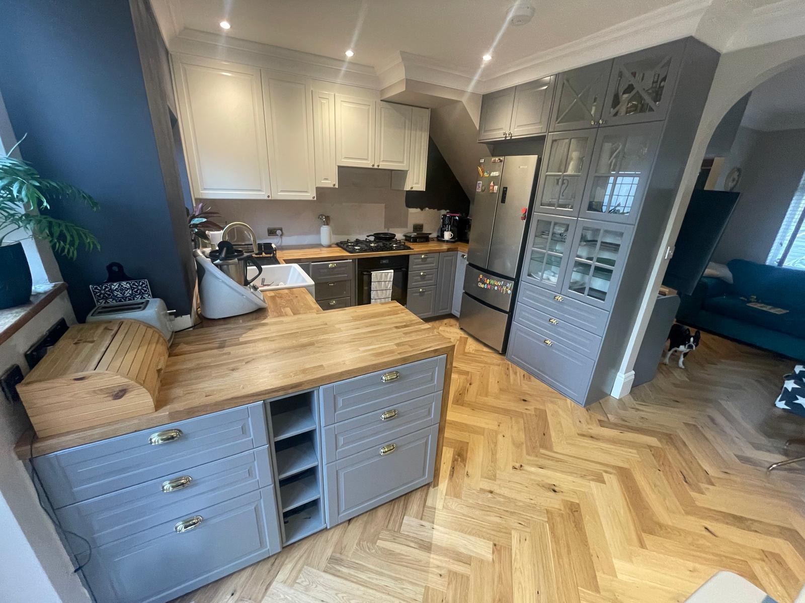 Jasna kuchnia z szarymi i białymi szafkami, drewnianym blatem, parkietem w jodełkę i widocznym psem rasy Boston Terrier w tle.