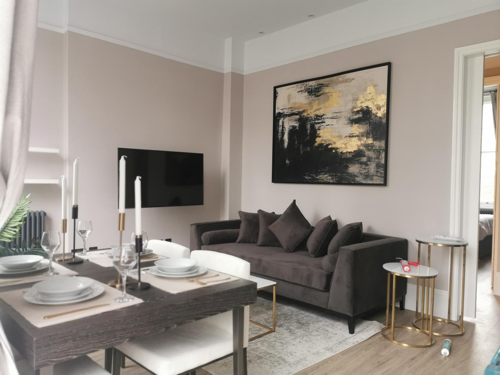 Jasne wnętrze salonu z ciemną sofą, abstrakcyjnym obrazem z elementami złota, stołem jadalnym z zastawą i telewizorem na ścianie, oświetlone światłem dziennym.