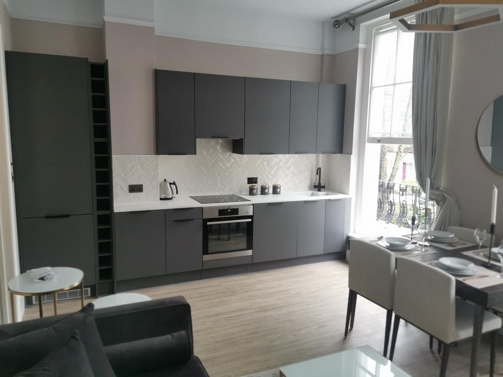 Nowoczesna kuchnia z szarymi szafkami, białym blatem i płytkami w jodełkę, widok na stół jadalny i okno z widokiem na zewnątrz.