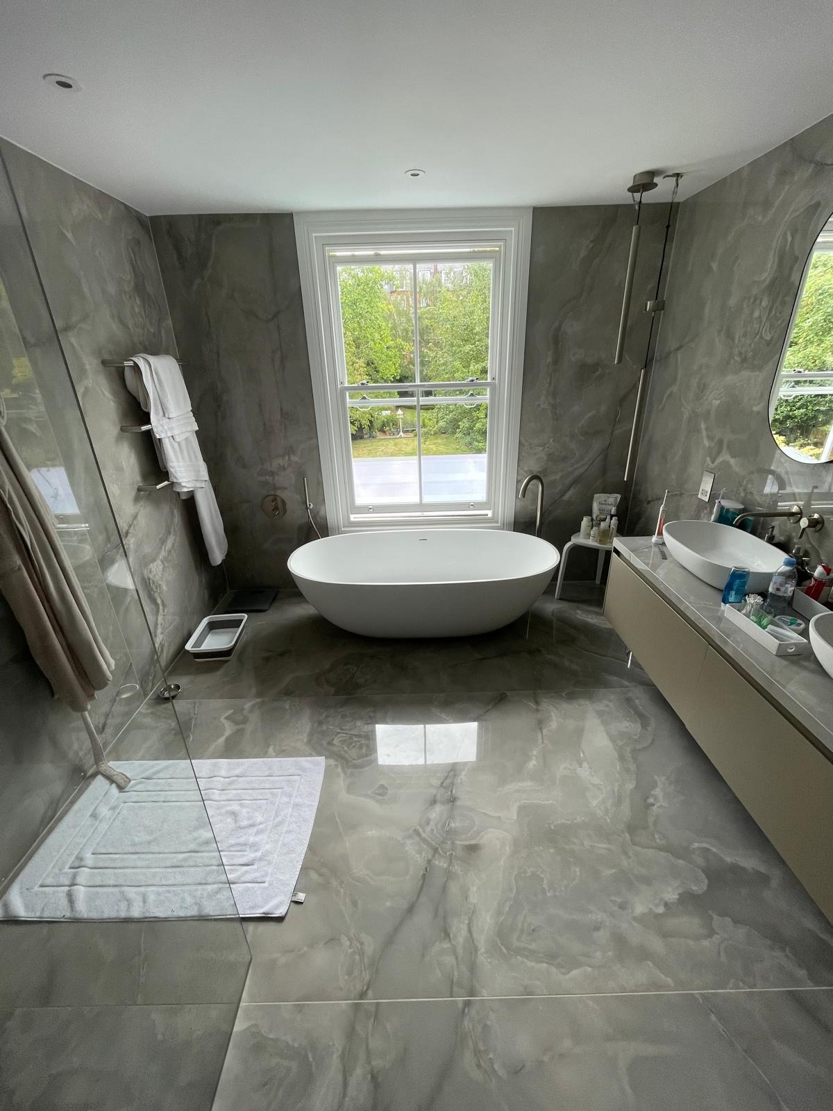 Nowoczesna łazienka z marmurowymi płytkami, wolnostojącą wanną przy oknie z widokiem na zieleń, szklanym panelem prysznicowym i podwójną umywalką na blacie.