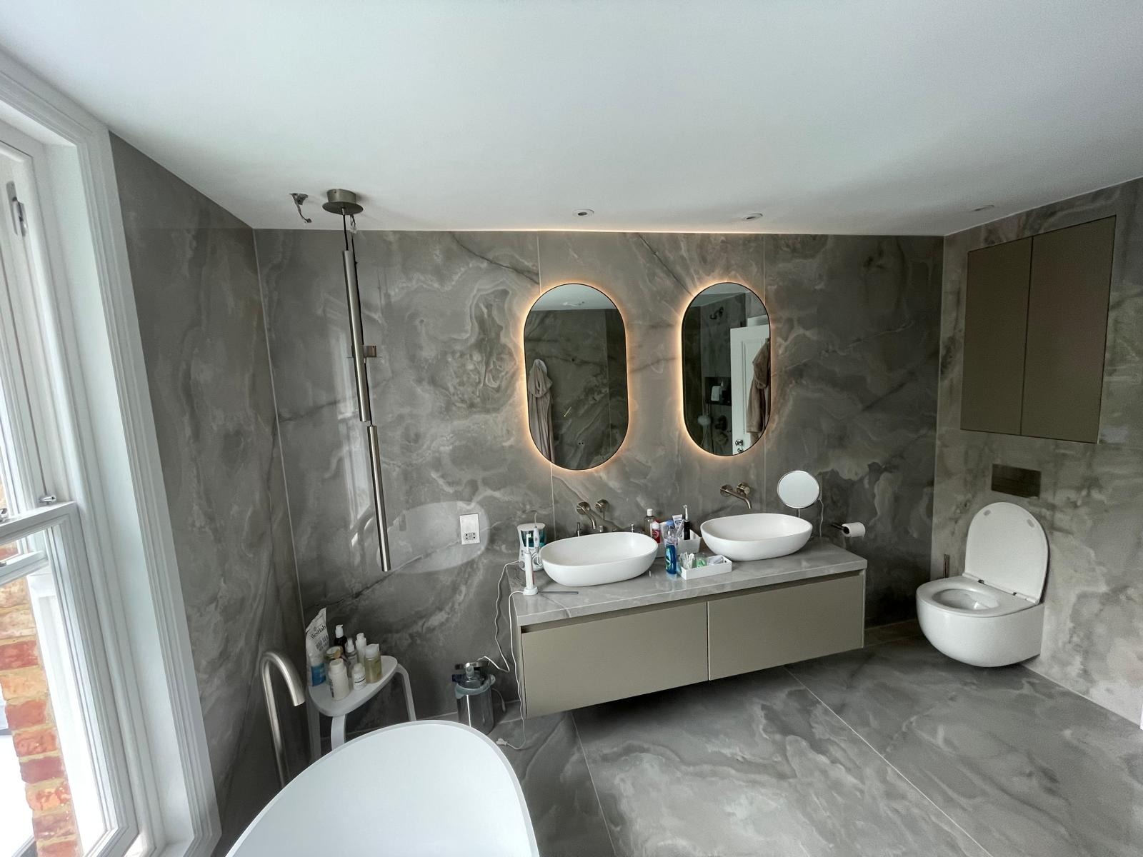 Nowoczesna łazienka wykończona szarymi płytkami imitującymi marmur, z podwójną umywalką nablatową, podświetlanymi lustrami owalnymi i wiszącą toaletą.