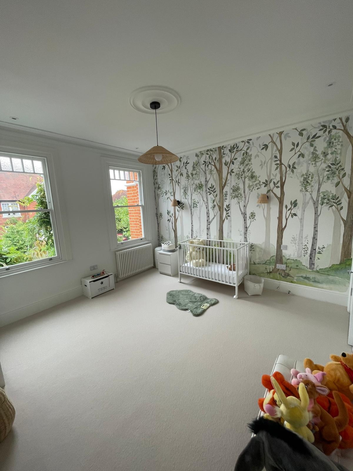 Jasny pokój dziecięcy z białymi ścianami, tapetą z motywem leśnym, białym łóżeczkiem i dywanikiem w kształcie misia, oświetlony lampą z abażurem z naturalnego materiału