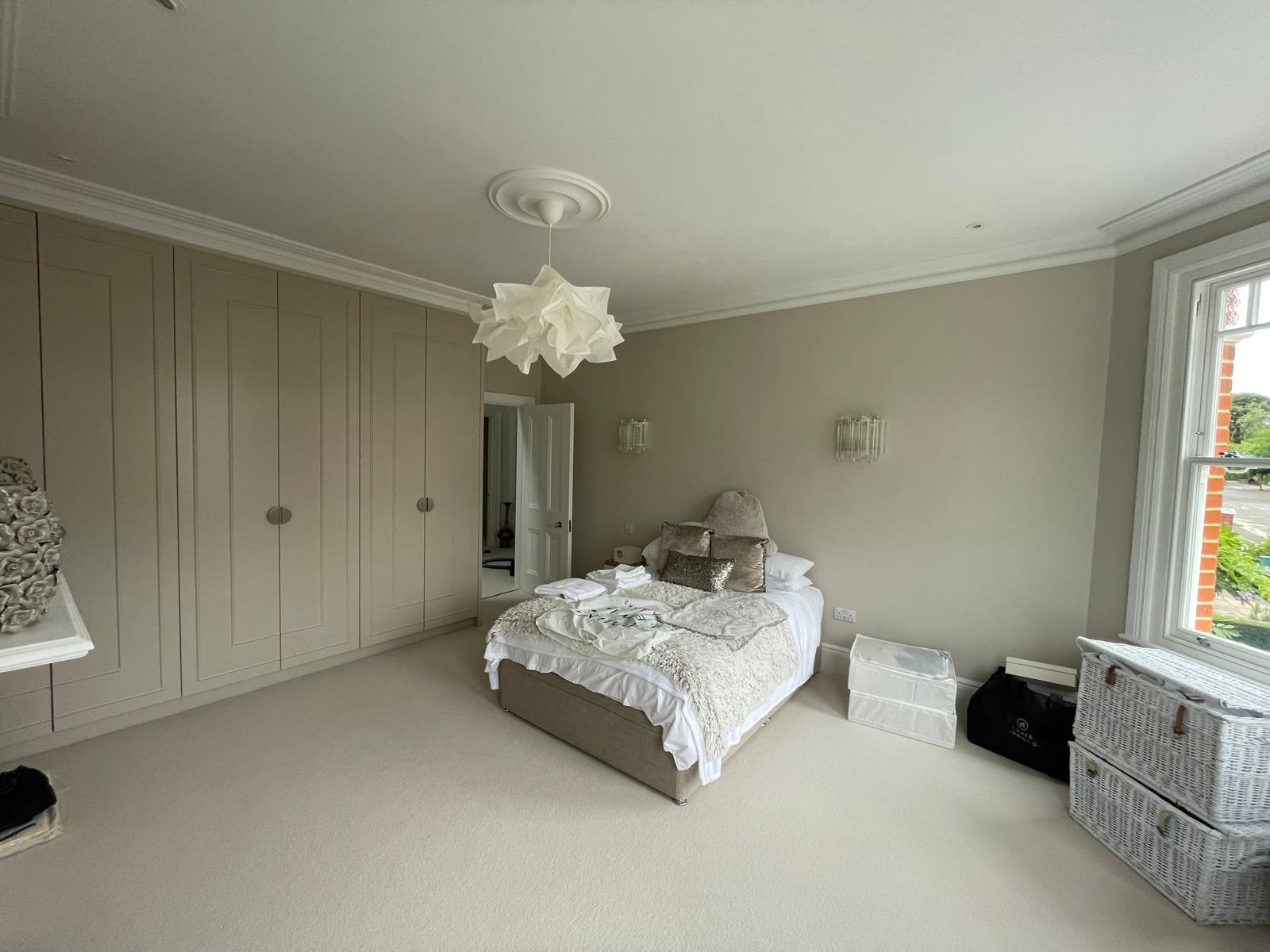 Neutralna sypialnia z beżowymi ścianami, zabudowaną szafą, teksturowanym dywanem i łóżkiem z dekoracyjnymi poduszkami oraz lampą w kształcie kwiatu.