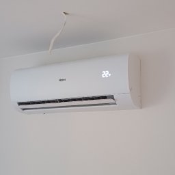 Adam Tarnacki-Instalacje - Biały klimatyzator Haier z widocznym wyświetlaczem temperatury 22°C, zamontowany na jasnej ścianie w pomieszczeniu, z fragmentem szafy po lewej stronie.