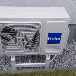 Adam Tarnacki-Instalacje - Biała jednostka zewnętrzna klimatyzacji Haier, zamontowana na elewacji budynku na niskim stelażu, otoczona kamieniami ozdobnymi.