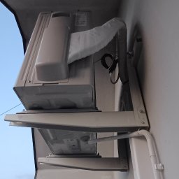 Adam Tarnacki-Instalacje - Zamontowana jednostka zewnętrzna klimatyzacji na wspornikach przy elewacji domu. Widoczne przewody i rury odprowadzające skropliny. Ujęcie z dołu, widoczne fragmenty dachu.