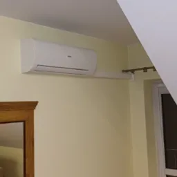 Biały klimatyzator Haier zamontowany na żółtej ścianie w pokoju z beżową sofą i oknem. Widoczny fragment ramy lustra.