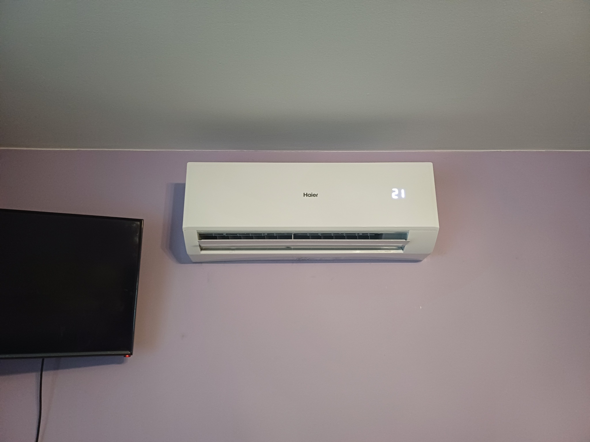 Biały klimatyzator marki Haier zamontowany na fioletowej ścianie w pomieszczeniu, obok widoczny fragment czarnego telewizora.