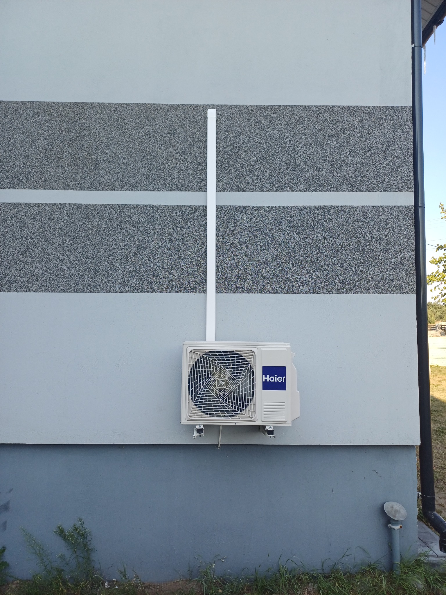 Biała jednostka zewnętrzna klimatyzatora Haier zamontowana na elewacji budynku z szarego tynku strukturalnego i gładkiego. Biała rura odprowadzająca skropliny biegnie pionowo w górę.