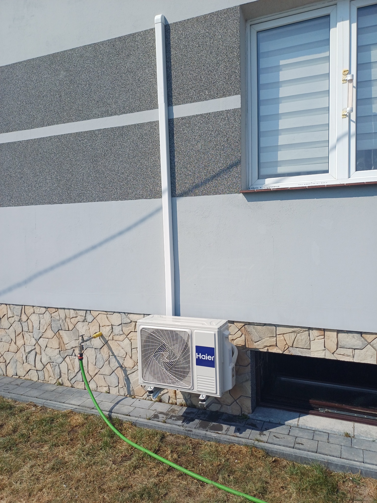 Biała jednostka zewnętrzna klimatyzatora Haier zamontowana na elewacji domu z dekoracyjnym kamieniem, widoczny zawór z podłączonym wężem ogrodowym.