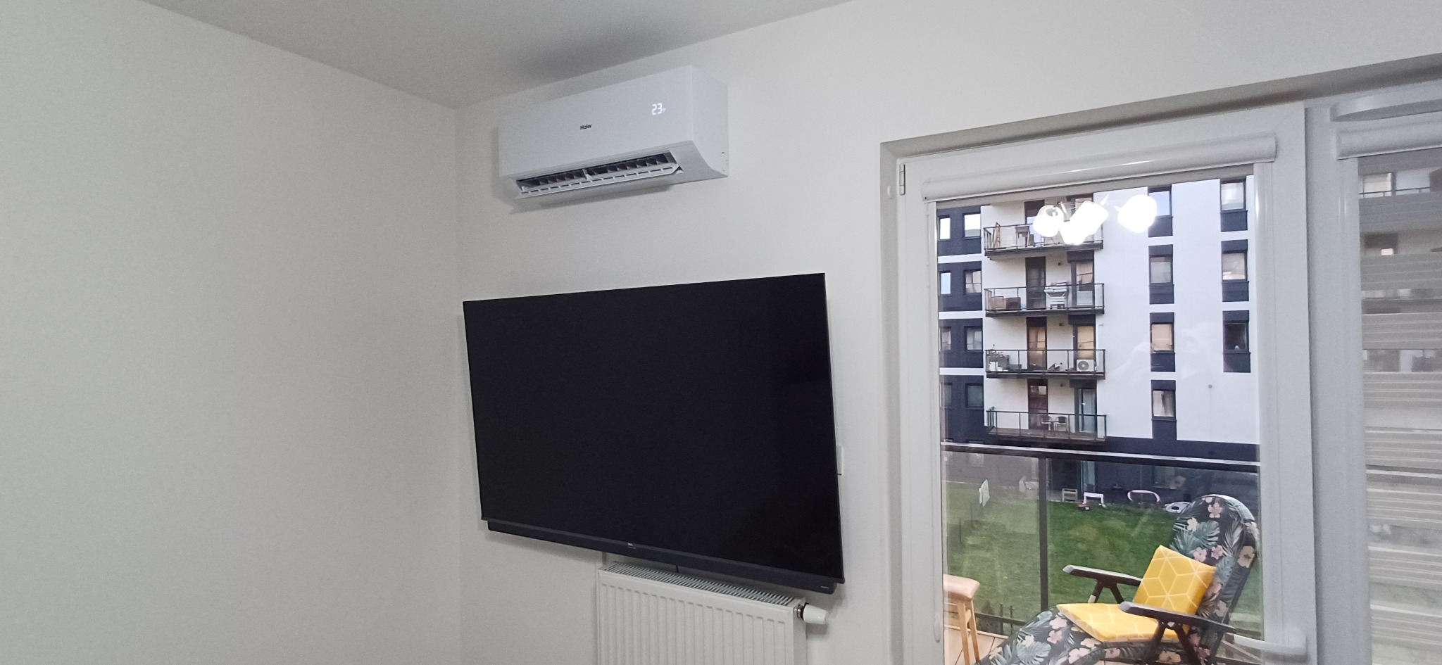 Biały klimatyzator Haier z wyświetlaczem temperatury 23 stopnie Celsjusza zamontowany nad czarnym telewizorem i białym grzejnikiem w jasnym pokoju z widokiem na balkon i budynek mieszkalny.