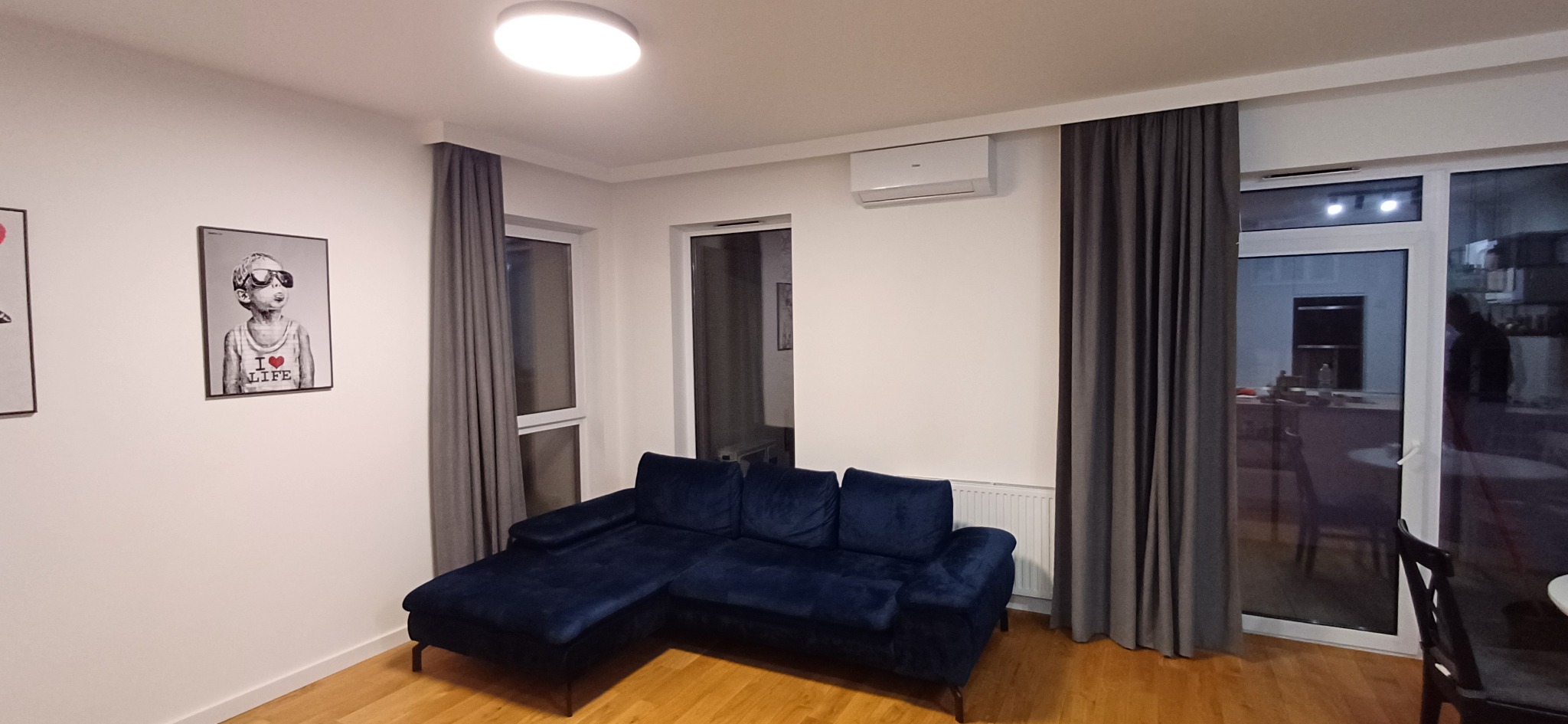 Nowoczesny salon z ciemnoniebieską kanapą narożną, klimatyzatorem na ścianie, szarymi zasłonami i plakatami na ścianie.